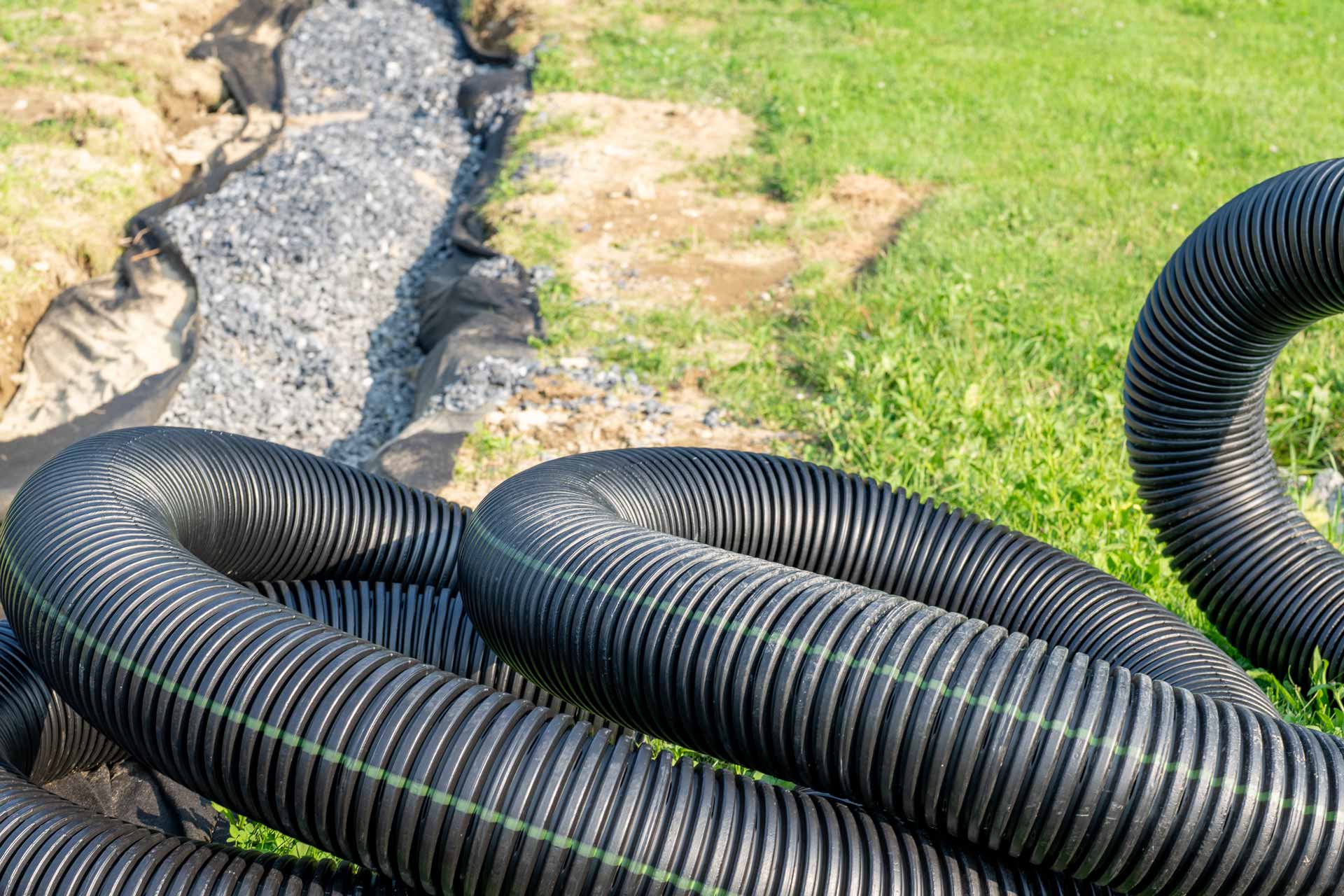 Corrugated pipes laying on the grass in front of a trench where a new French drain is being installed